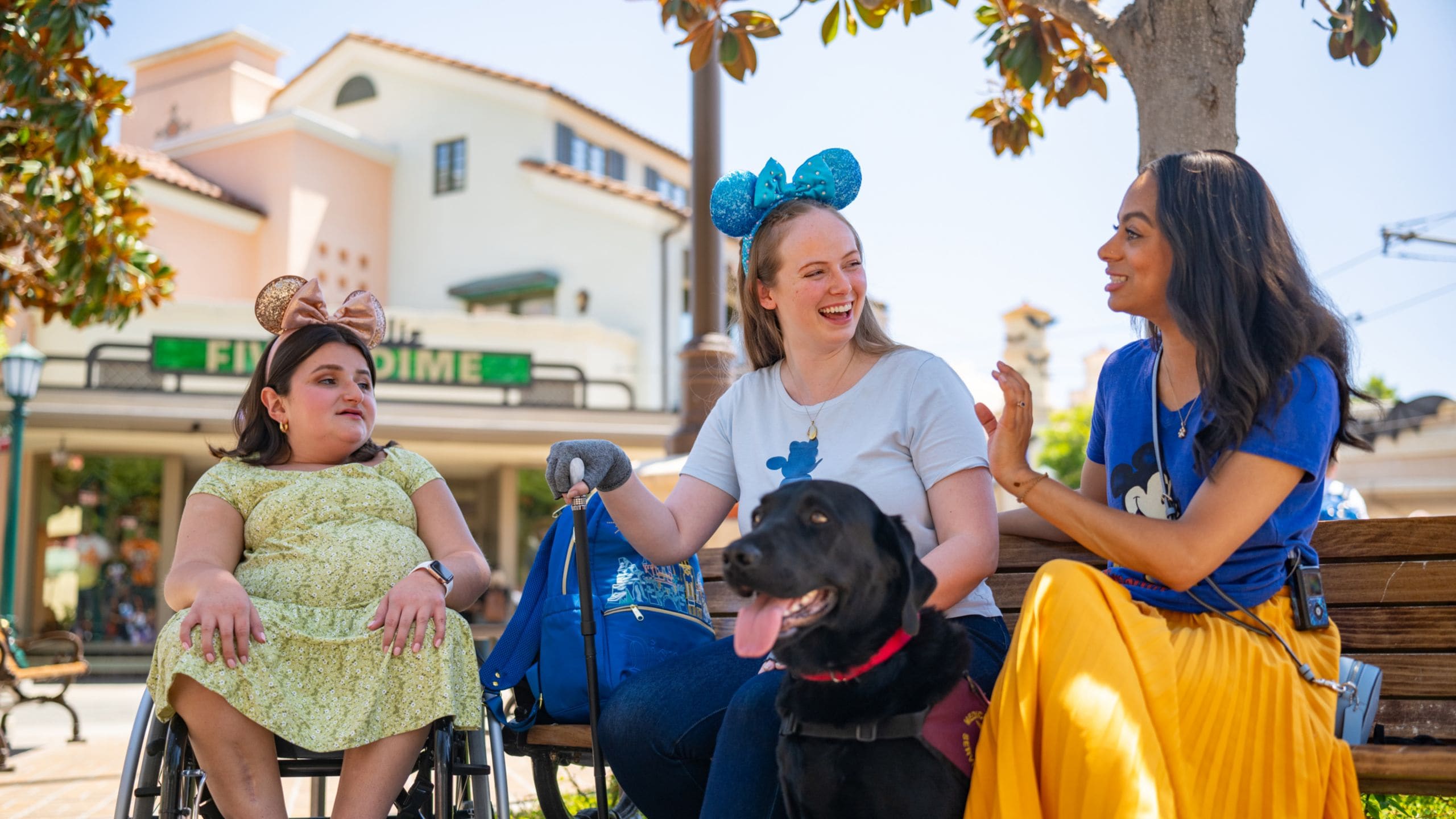 A woman in a wheelchair, a woman with a cane, and a woman with a service dog are engaged in conversation while sitting outside in Disney California Adventure Park.