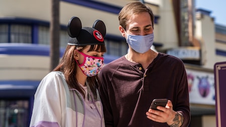 Un Visitante con una máscara facial y orejas de Mickey y otro Visitante con una máscara mira un teléfono celular