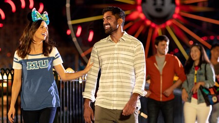 Dos parejas caminando por Pixar Pier de noche con las luces de Pixar Pal A Round destellando en el fondo
