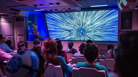 Dentro de Star Tours en Disneyland Park, los Visitantes sentados ven los efectos de luz apoderándose de la pantalla, mientras C 3 P O se encuentra de pie a un costado 