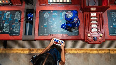 Una niña controla su nuevo droide serie R sobre una consola de taller con planos de construcción de droides.