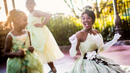 Princesa Tiana, em um pátio cercado, sorri com 2 meninas vestidas com fantasias da Princesa Tiana