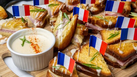 A board set with miniature Croque Monsieur sandwiches decorated with French flags and a small bowl of béchamel sauce