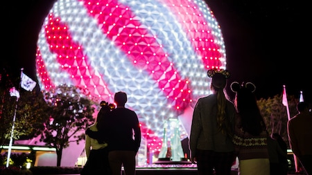 Epcot Guests look up at Spaceship Earth, which is lit up with pink and white diagonal stripes