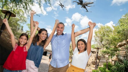 A man, woman and 2 daughters smiling for a photo in Star Wars Galaxy’s Edge, while starfighters appear in the sky above