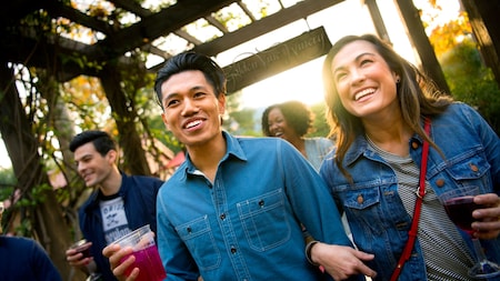 A young couple holding drinks as they walk beneath a pergola
