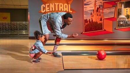 A dad teaches his daughter how to roll a bowling ball down an alley