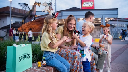 A family of 5 sit along a low wall outside of The Lego Store in Disney Springs