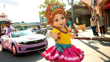 Fancy Nancy waving as she walks in the cavalcade, followed by a cars with Doc McStuffins and Vampirina waving