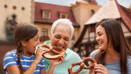 Grandfather bites playfully into a huge pretzel his granddaughter offers, while big sister laughs