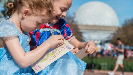 2 young Guests in princess attire, drawing on maps of Epcot with Spaceship Earth in the background