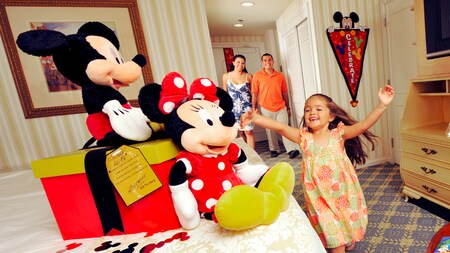 An excited young girl and her parents entering a hotel room to discover Mickey and Minnie Mouse plush dolls and a gift box on the bed