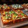 Several pans of Detroit style pizza, featuring one with jalapenos, cilantro, pepperoni and crumbled cheese