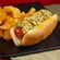 A large hot dog drizzled with condiments in a bun, sitting next to a serving of crisp potato chips