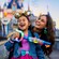 A girl wearing a Disneyland 70th anniversary ear headband and her mother, both playing with a bubble blower near Sleeping Beauty Castle