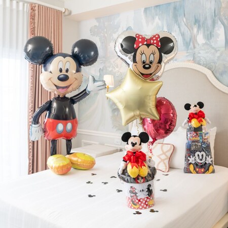 Un globo grande de aluminio con forma de Mickey Mouse, junto a otros globos y dos cestas de regalos con productos de Mickey Mouse que incluyen peluches, sombreros de orejas y un bolso con cordón en una cama de hotel