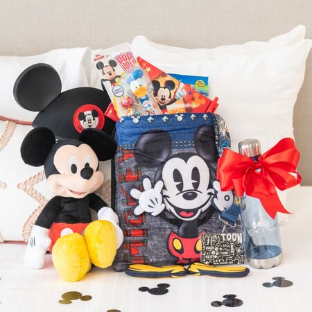 Un peluche de Mickey Mouse, un sombrero de orejas y un bolso con cordón en una cama de hotel junto a una botella de Disney, una cinta, blocs de notas y bolígrafos