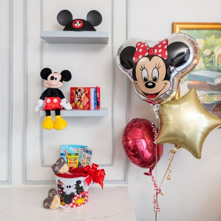 Un estante de una suite de hotel con un sombrero de orejas de Mickey Mouse, un peluche y un bloc de notas junto a un montón de globos y una cesta de regalos con dulces y un libro para colorear