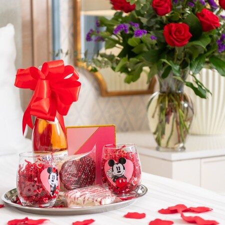 Una bandeja con copas de Mickey y Minnie Mouse, una botella de vino, dulces de arroz inflado y pétalos de rosa en una cama junto a un florero con flores y rosas