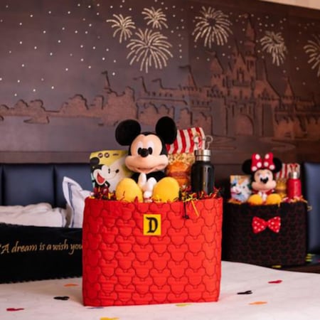A Mickey Mouse plush in a gift box sit on a hotel bed