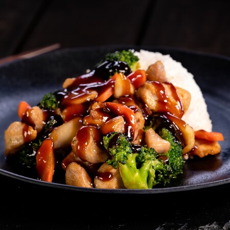 Un plato de pollo teriyaki con vegetales salteados y arroz al vapor sobre un individual de pizarra con palillos chinos a un costado