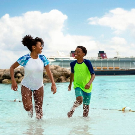 A boy and girl run along the shore on Disney Castaway Cay with a Disney Cruise Line ship in the background