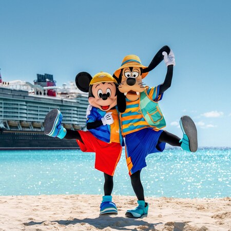 Mickey Mouse e Goofy em traje de banho posando na praia perto de um navio de cruzeiro da Disney atracado
