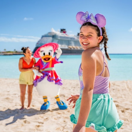 A child smiling as she looks over her shoulder while Daisy Duck stands arm and arm with a Guest on the beach in the background