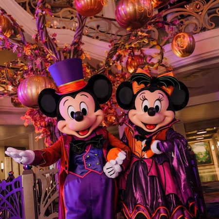 Mickey Mouse and Minnie Mouse in Halloween costumes beneath a tree decorated with pumpkins