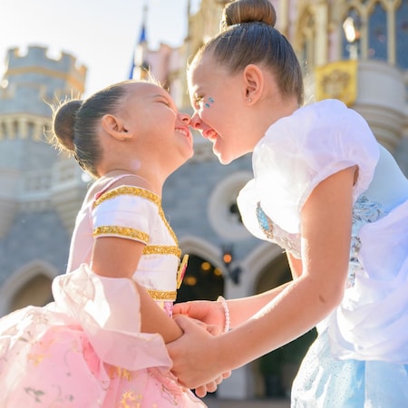 Two girls dressed as Belle and Cinderella touching noses in front of Sleeping Beauty Castle