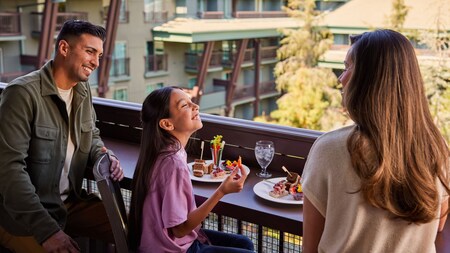 A family of 3 dining on a balcony overlooking a courtyard in Disneyland Hotel