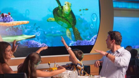 A family of 4 watching an animated sea turtle on a digital screen while dining at Animator's Palate on a Disney cruise ship