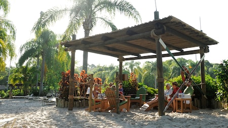 Guests sit on a beach in lounge chairs inside an open-air, wooden shack