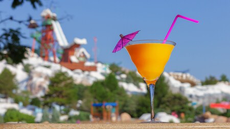 A cocktail with a straw and umbrella on a table in front of a snowy mountain with a waterslide