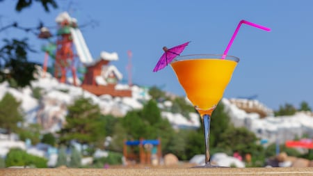 A cocktail with a straw and umbrella on a table in front of a snowy mountain with a waterslide