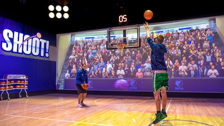 A virtual crowd and an NBA Experience employee watch a male Guest aim a basketball at a hoop in a basketball court setting
