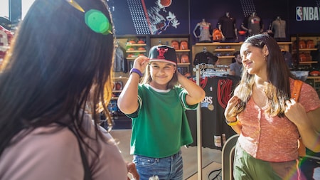 A young female Guest tries on a cap in front of 2 female friends inside the NBA Store