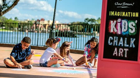 A group of kids sit on the ground drawing with chalk near a sign that reads ‘Imagination Kids’ Chalk Art’