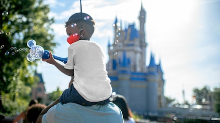 Little boy with a bubble wand sits on dads shoulders with Cinderellas Castle in the background