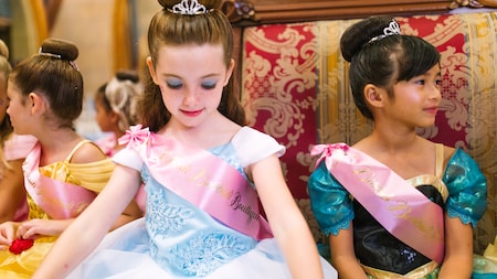 Young girls wearing princess gowns, sashes and crowns sit together