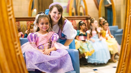 A young girl in a princess gown and her Fairy Godmother in Training smile at the mirror