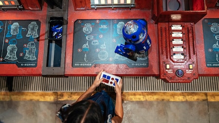 A young girl controls her new R series droid atop a workshop console featuring construction blueprints of droids