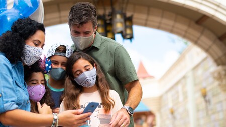 Family stands together in Disney's Magic Kingdom Park with face coverings looking at cell phone for planning purposes