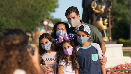 A family stands together wearing face coverings in front of partner's statue at Disney World while PhotoPass takes their photo