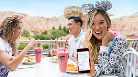 A teen Guest demonstrates a food order on a mobile device while her friends eat in the background