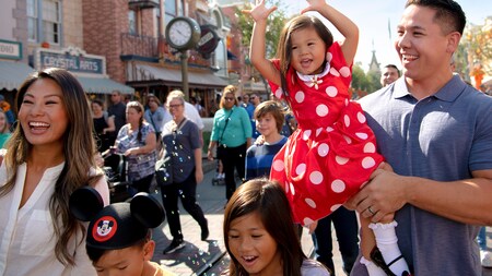 A young girl in a Minnie Mouse dress is carried by her father as their family walks down  Main Street USA
