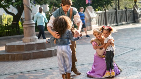 Two actors dressed as Rapunzel and Flynn Rider hug 2 young girls as other Guests pass by in the background