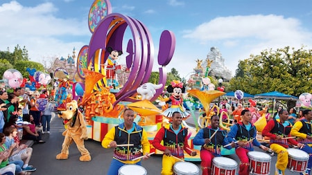 Drummers play snares in front of a parade float with Mickey Mouse, as Goofy interacts with the crowd
