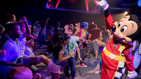 A large group of kids sing and dance along with Mickey, who is dressed in his Roadster Racers costume