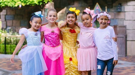 With their arms wrapped around each other, 5 young girls pose outside Sleeping Beauty Castle in princess costumes and accessories.